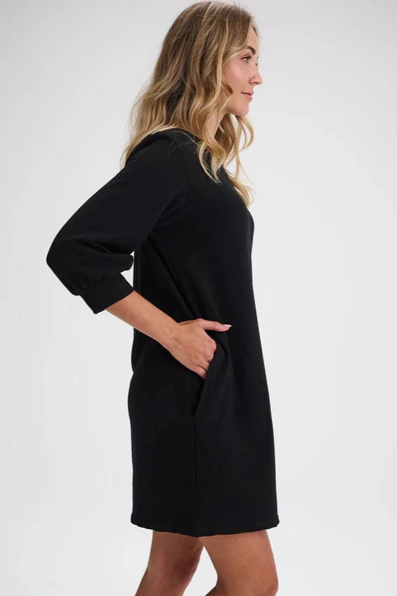 Side view of a woman wearing the Stukely Dress by Message Factory in Black, a semi-fitted bamboo/organic cotton fleece dress with a boat neck, 3/4 sleeves, side pockets, and a mid-thigh length. She is standing in front of a beige background. 