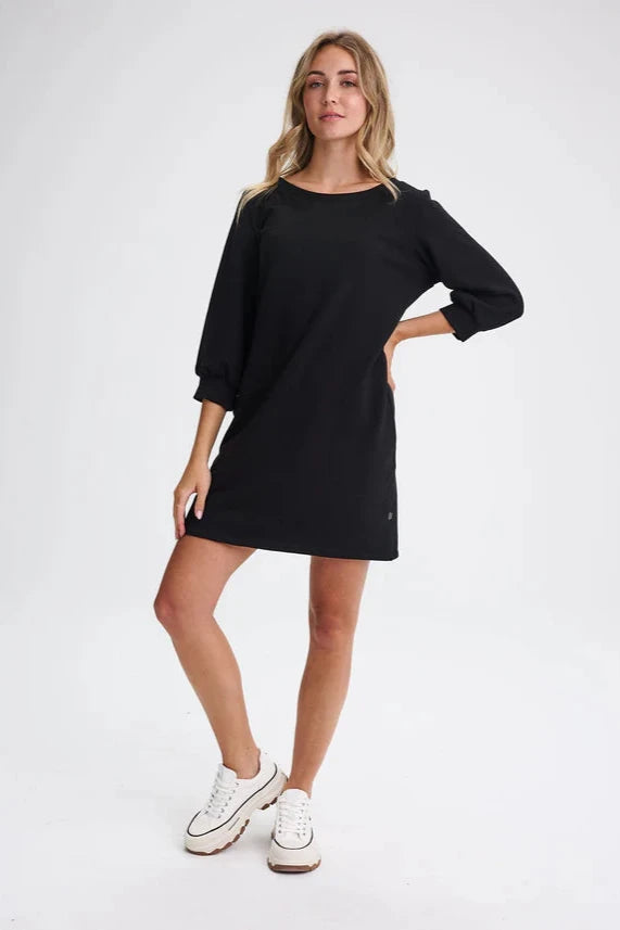 A woman wearing the Stukely Dress by Message Factory in Black, a semi-fitted bamboo/organic cotton fleece dress with a boat neck, 3/4 sleeves, side pockets, and a mid-thigh length. She is standing in front of a beige background. 