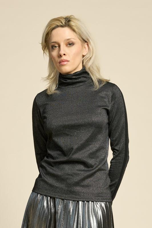A woman wearing the Mortel Top by Cokluch in Facet Black, a metallic turtleneck with long sleeves. She is wearing it with the Feyer Skirt in Glow Knit Silver and standing in front of a beige background. 
