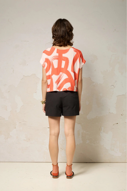 Back view of a woman wearing the Nyx top from Cockluch in Red Shodo, a short sleeved, loose fitting top with a drawstring detail at the bottom hem. She is wearing it with black shorts and standing in front of a textured background. 