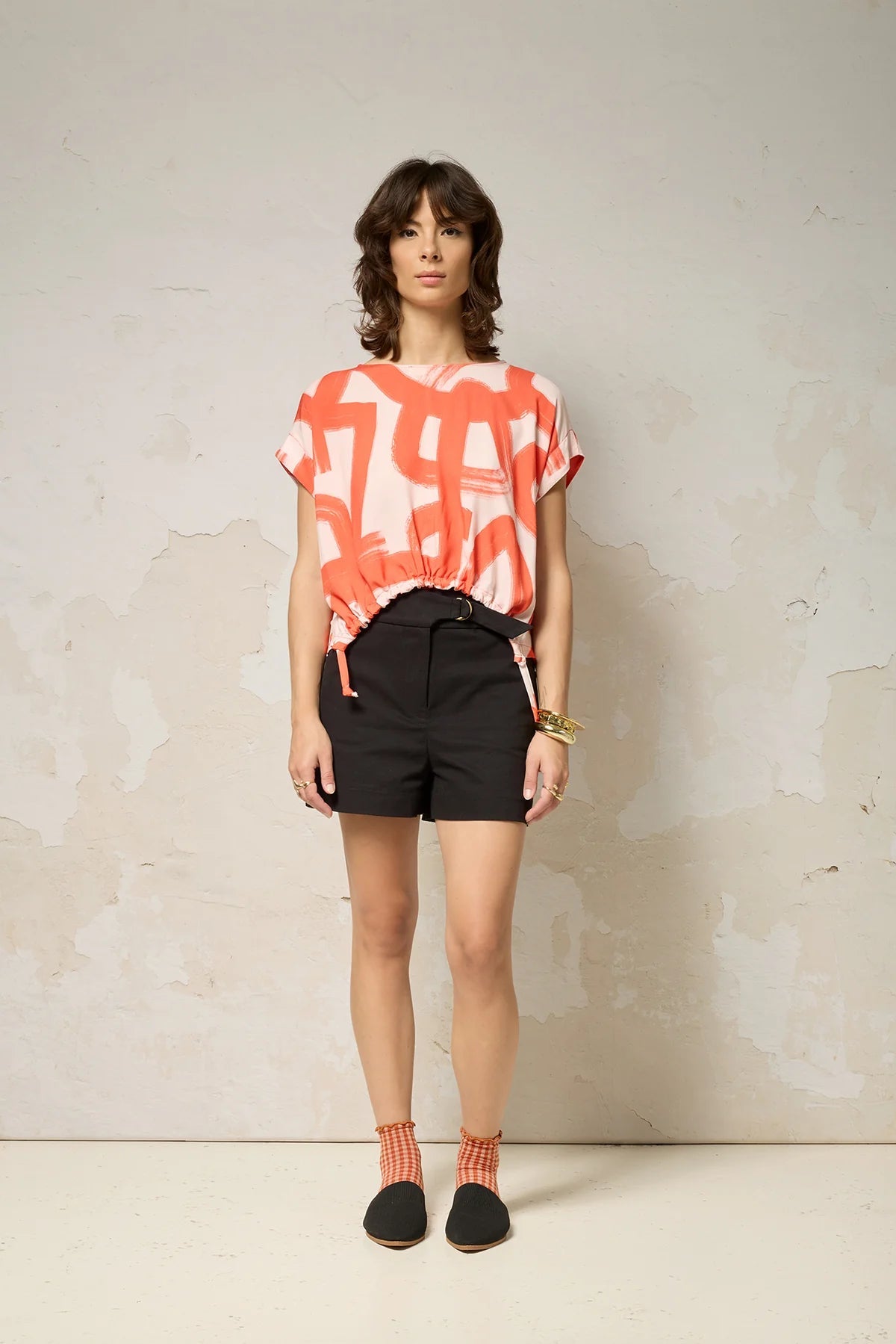 A woman wearing the Nyx top from Cockluch in Red Shodo, a short sleeved, loose fitting top with a drawstring detail at the bottom hem. She is wearing it with black shorts and standing in front of a textured background. 