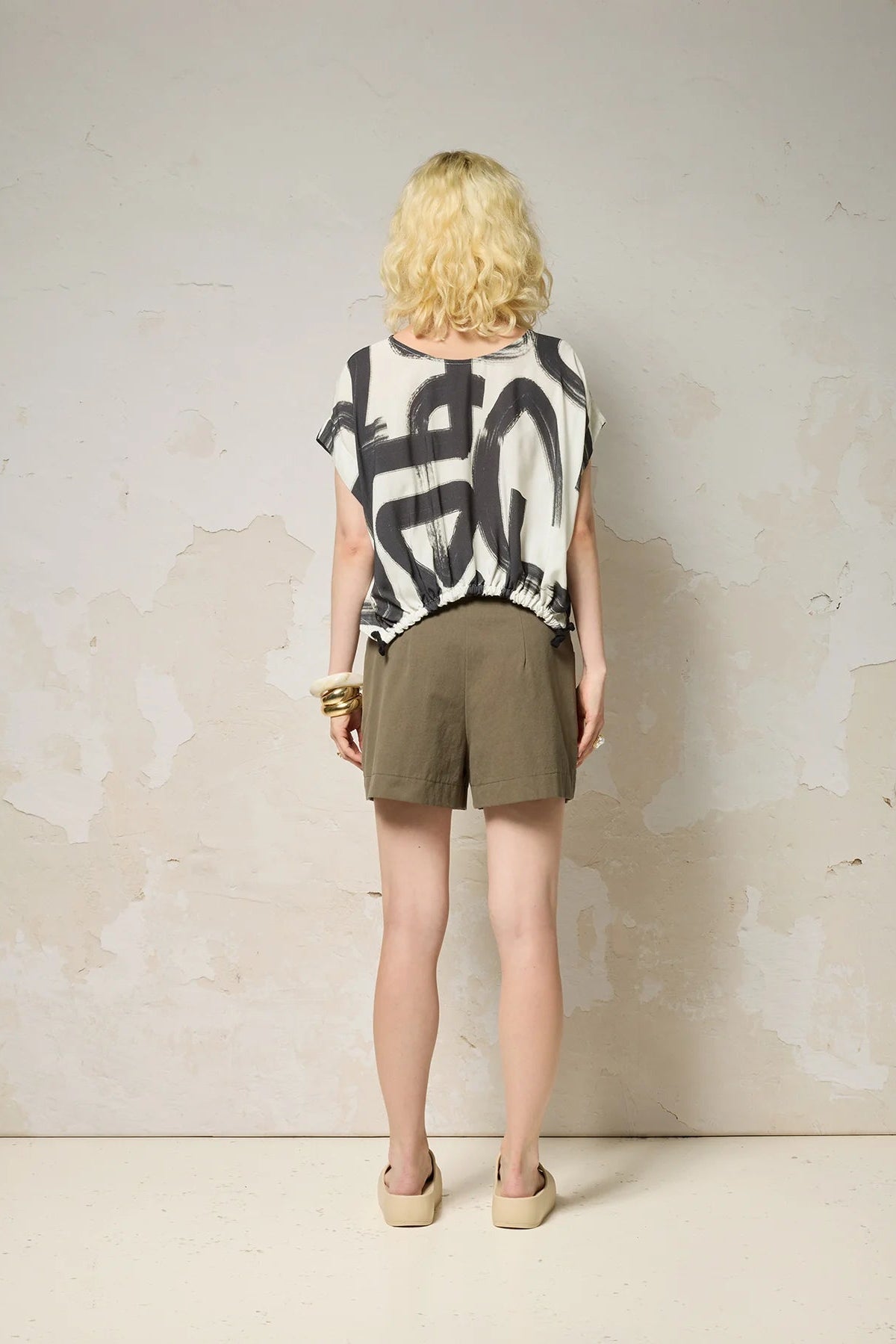 Back view of a woman wearing the Nyx top from Cockluch in Black Shodo, a short sleeved, loose fitting top with a drawstring detail at the bottom hem. She is wearing it with khaki shorts and standing in front of a textured background. 