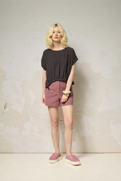 Waist up view of a woman wearing the Nyx top from Cockluch in Iron, a short sleeved, loose fitting top with a drawstring detail at the bottom hem. She is wearing it with pink shorts and standing in front of a textured background. 