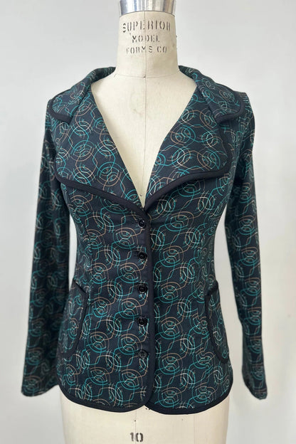 The Nara Jacket by Kollontai in Turquoise, a blazer with a turquoise on black geometric print, with a rounded lapel collar, slim fit, and side pockets, is shown on a mannequin in front of a grey background. 