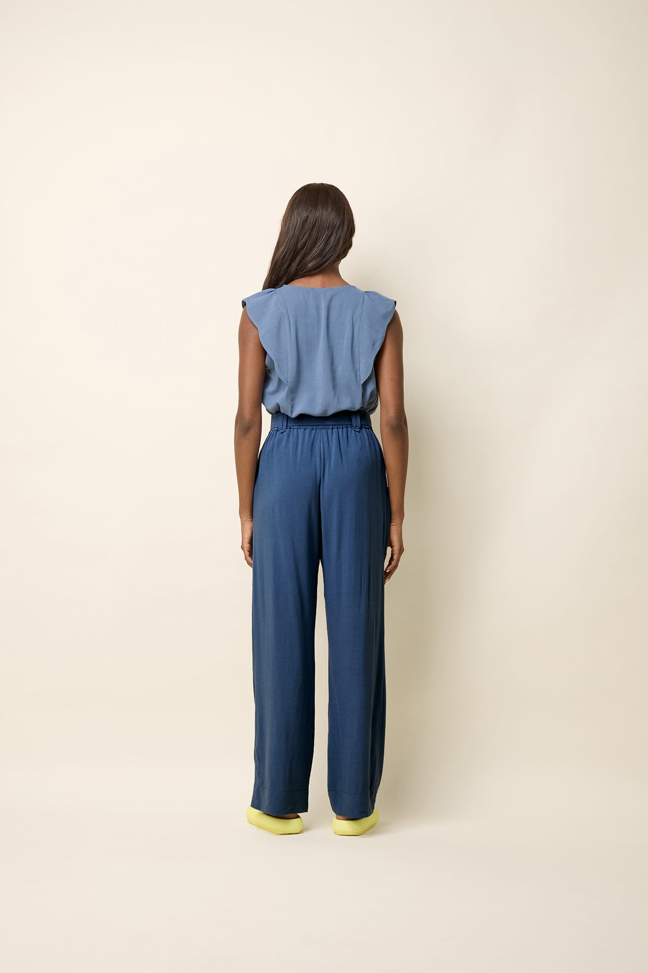 Back view of a woman wearing the Neema Top by Cokluch in Ocean, a light sleeveless top with ruffle details at the shoulders, a round neck, and a mid-hip length. She is wearing it with blue pants and standing in front of a beige background. 