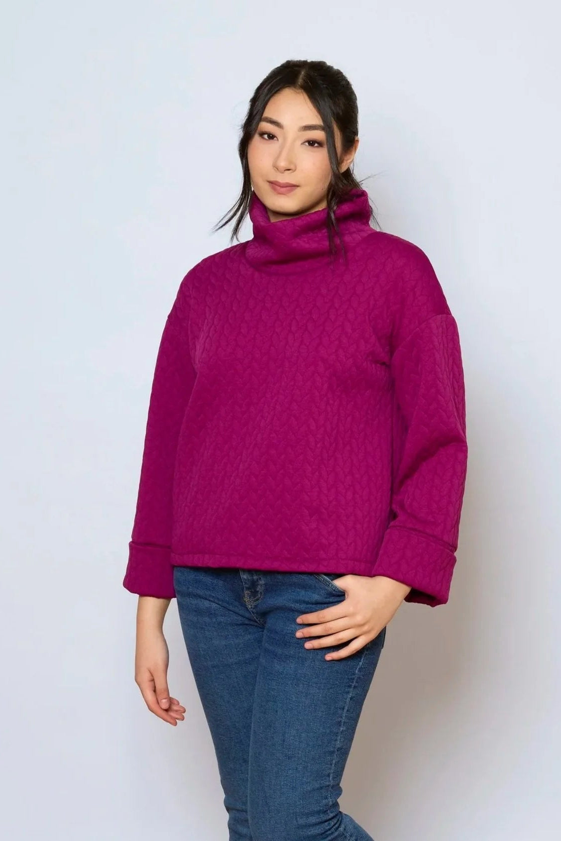 A woman wearing the Nori Top by Tangente in Magenta, a relaxed top in quilted fabric with a large cowl neck and cuffed drop sleeves. She is wearing it with jeans and standing in front of a white background. 