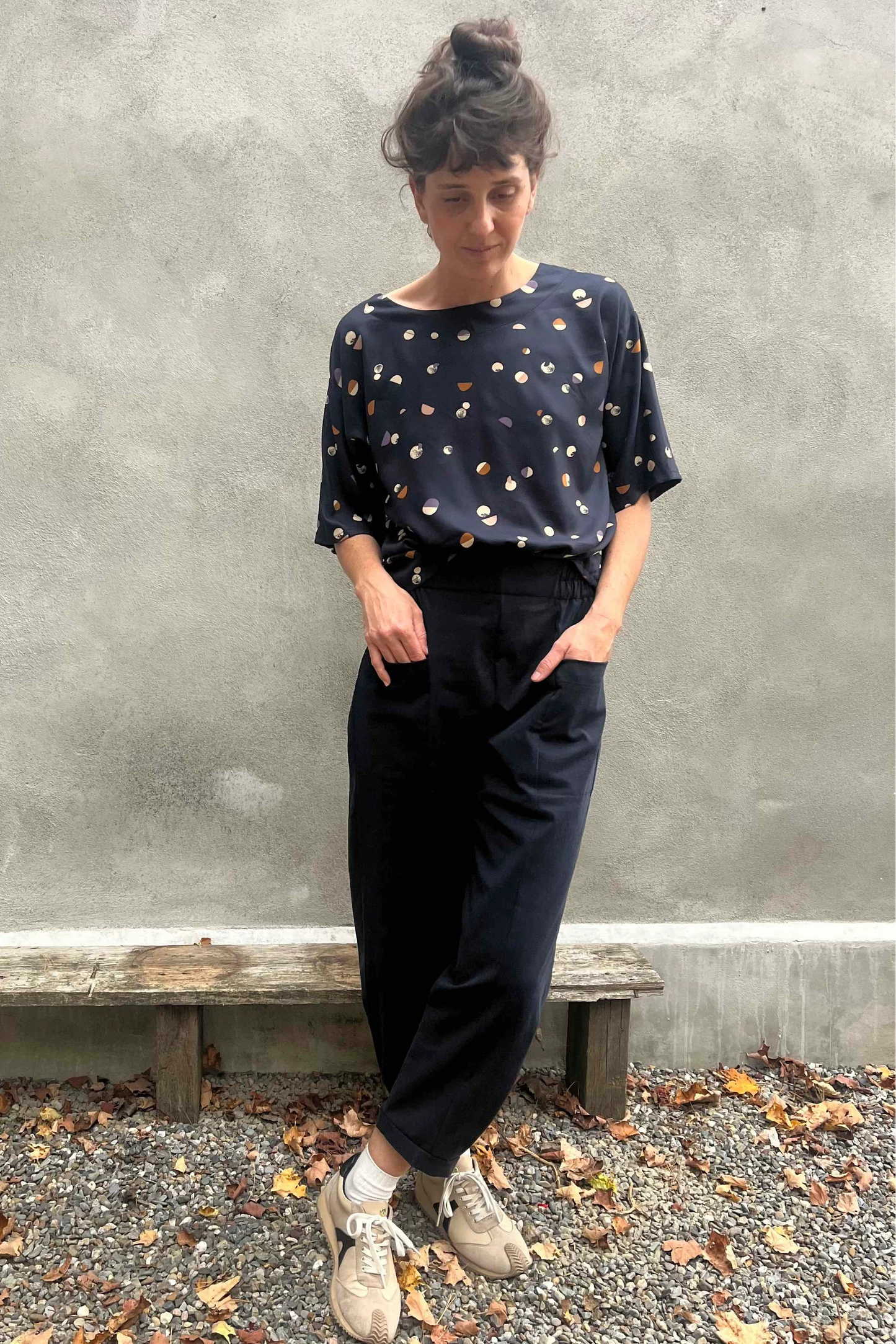 A woman wearing the Nyssa Pants by Kazak in Navy, barrel-legged pants with an elastic waistband and patch pockets. She is wearing them with the Abelia Top in Midnight Moons and standing outside by a grey wall. 
