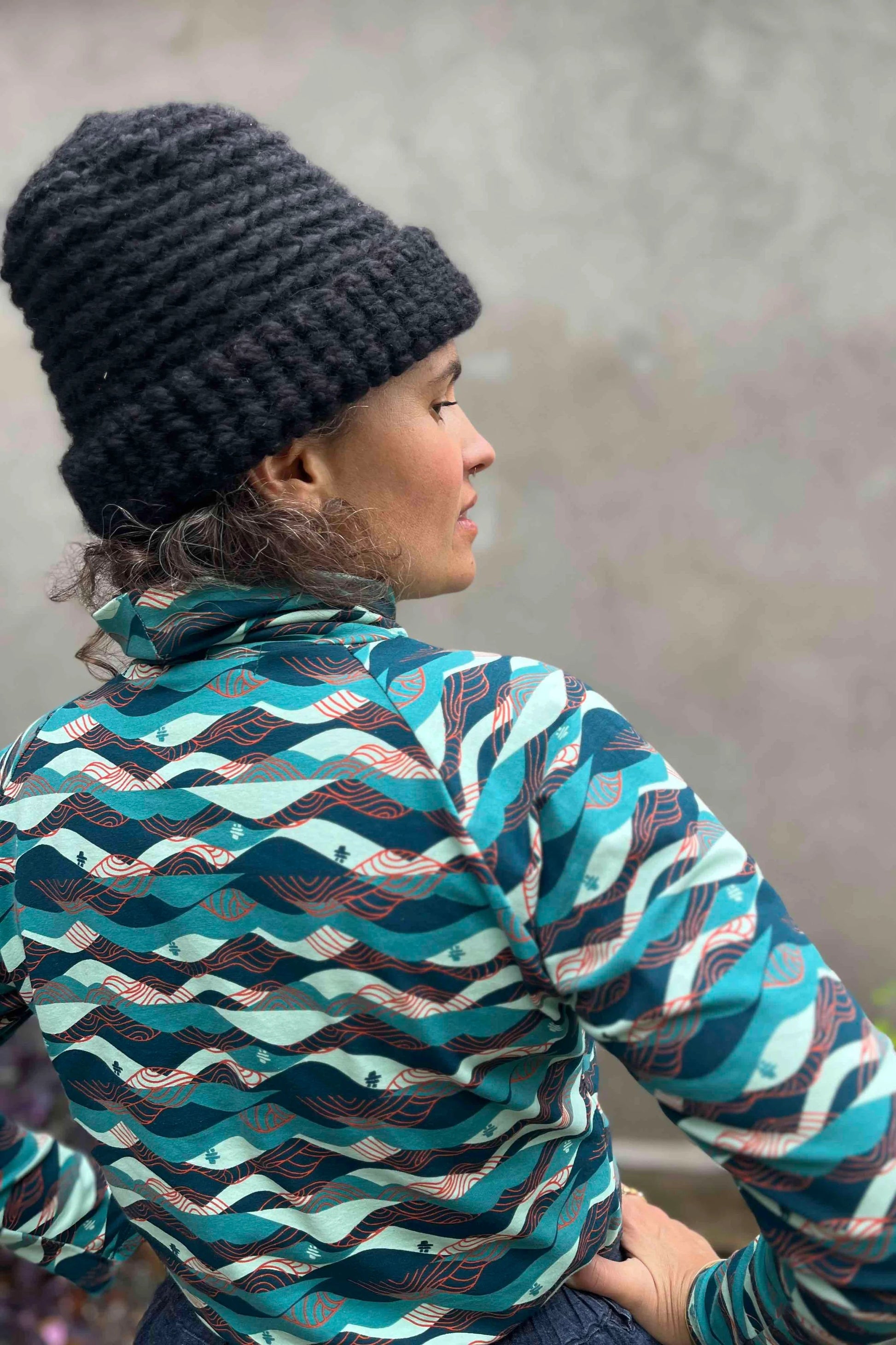 A woman wearing the Oris Raglan Turtleneck by Kazak in Inuksuk print, with a pattern of rolling waves and tiny inukshuks in shades of blue. 