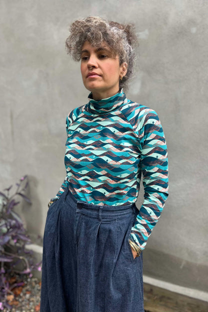 A woman wearing the Oris Raglan Turtleneck by Kazak in Inuksuk print, with a pattern of rolling waves and tiny inukshuks in shades of blue. She is wearing it with jeans and standing outside. 