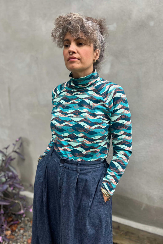 A woman wearing the Oris Raglan Turtleneck by Kazak in Inuksuk print, with a pattern of rolling waves and tiny inukshuks in shades of blue. She is wearing it with jeans and standing outside. 