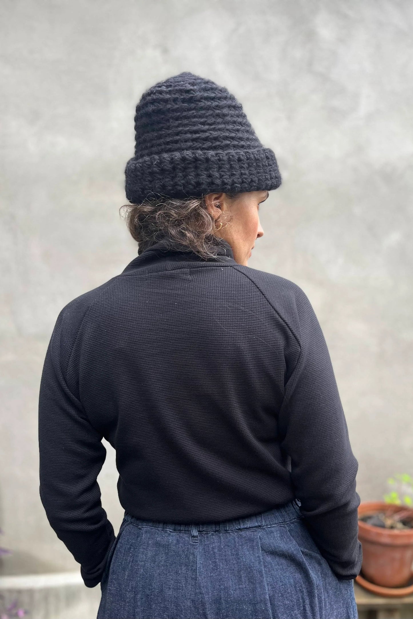 Back view of a woman wearing the Oris Raglan Turtleneck by Kazak in Black waffle. She is wearing it with jeans and standing outside. 
