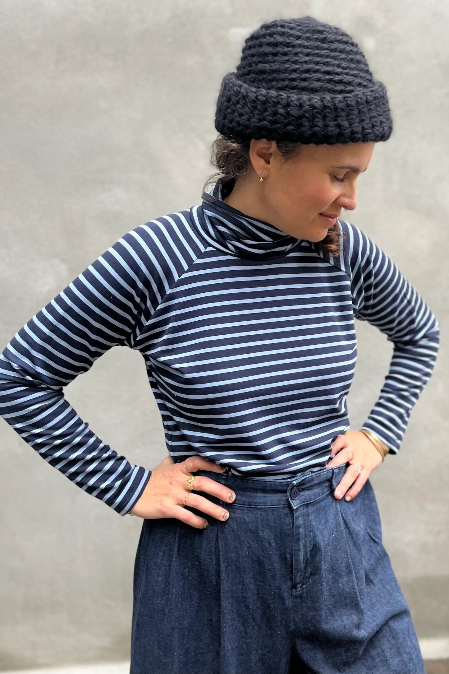 A woman wearing the Oris Raglan Turtleneck by Kazak in Blue Stripe. She is wearing it with jeans and standing outside. 