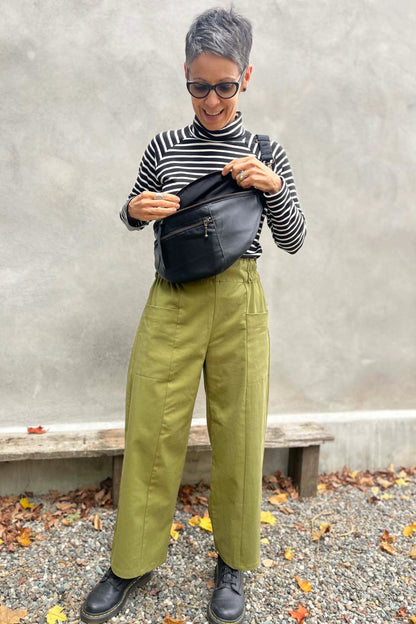 A woman wearing the Olea Pants by 
Kazak in Olive, wide-legged ankle-length pants with a flat-front elastic waist and large patch pockets. She is wearing them with the Oris Top in Blue Stripe and standing outside by a grey wall. 
