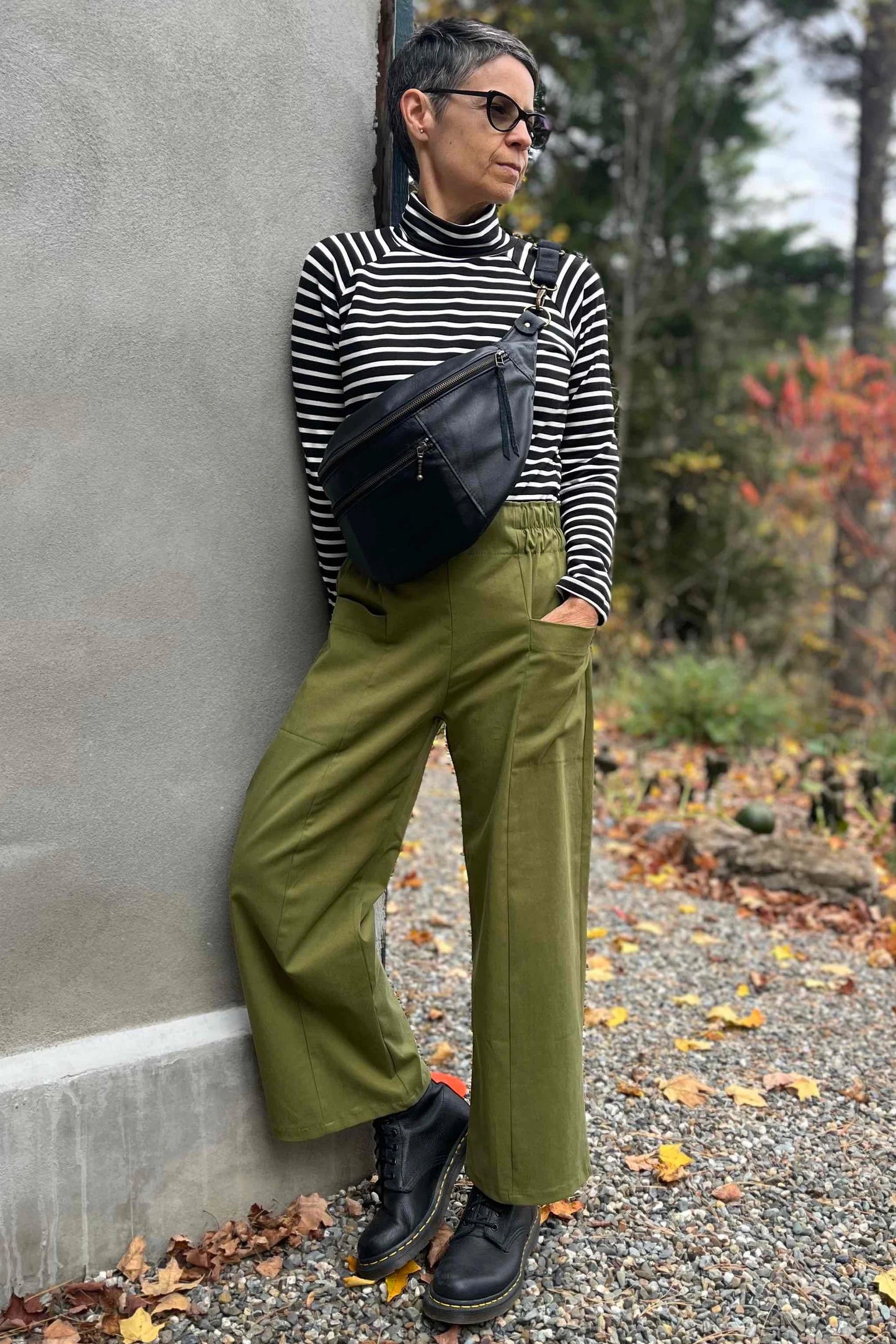 A woman wearing the Olea Pants by 
Kazak in Olive, wide-legged ankle-length pants with a flat-front elastic waist and large patch pockets. She is wearing them with the Oris Top in Blue Stripe and standing outside by a grey wall. 