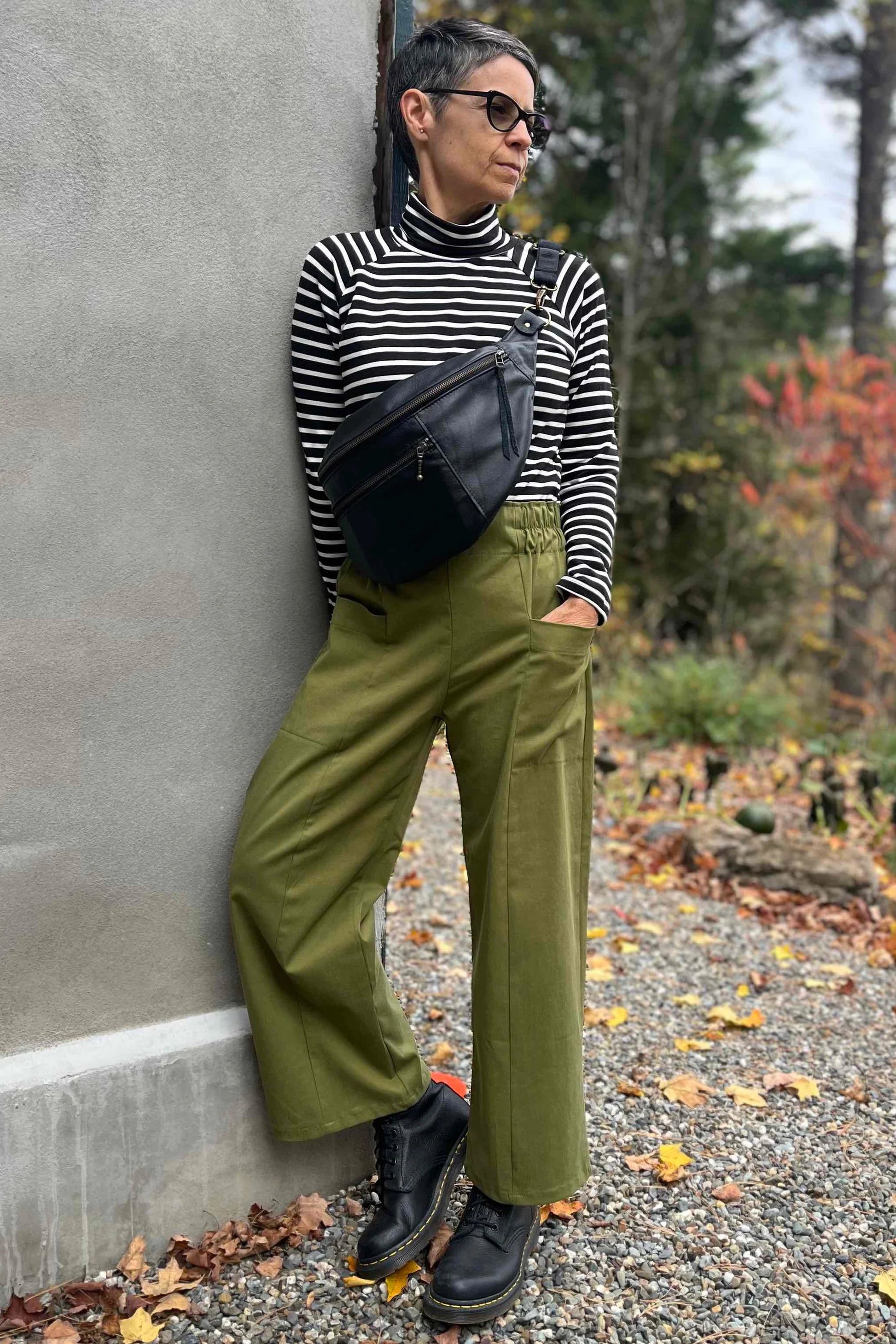 A woman wearing the Olea Pants by 
Kazak in Olive, wide-legged ankle-length pants with a flat-front elastic waist and large patch pockets. She is wearing them with the Oris Top in Blue Stripe and standing outside by a grey wall. 
