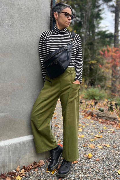 A woman wearing the Olea Pants by 
Kazak in Olive, wide-legged ankle-length pants with a flat-front elastic waist and large patch pockets. She is wearing them with the Oris Top in Blue Stripe and standing outside by a grey wall. 