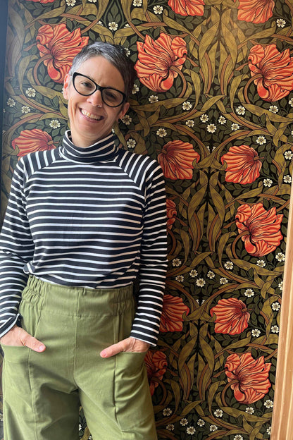 A woman wearing the Olea Pants by 
Kazak in Olive, wide-legged ankle-length pants with a flat-front elastic waist and large patch pockets. She is wearing them with the Oris Top in Blue Stripe and standing in front of a floral wallpapered wall. 