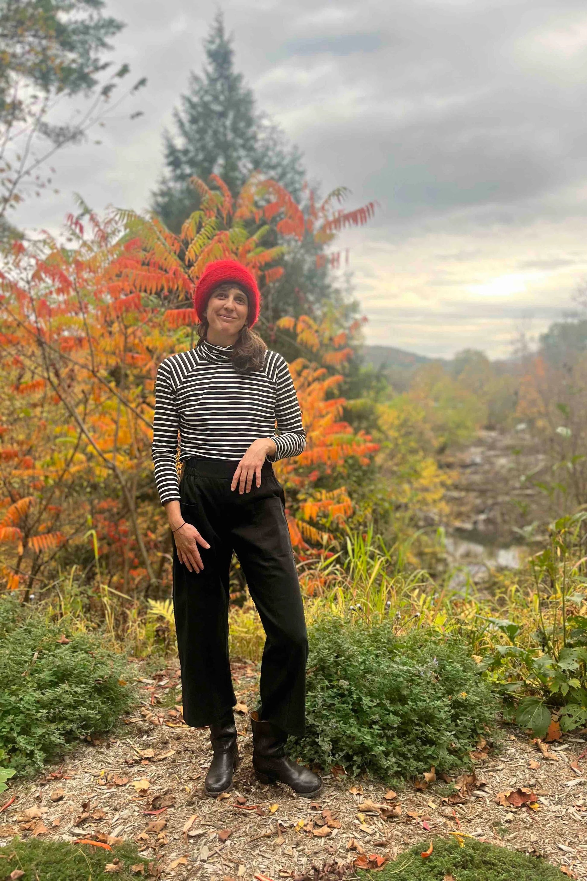 A woman wearing the Olea Pants by 
Kazak in Black, wide-legged ankle-length pants with a flat-front elastic waist and large patch pockets. She is wearing them with the Oris Turtleneck in Black Stripe and standing in a forest.