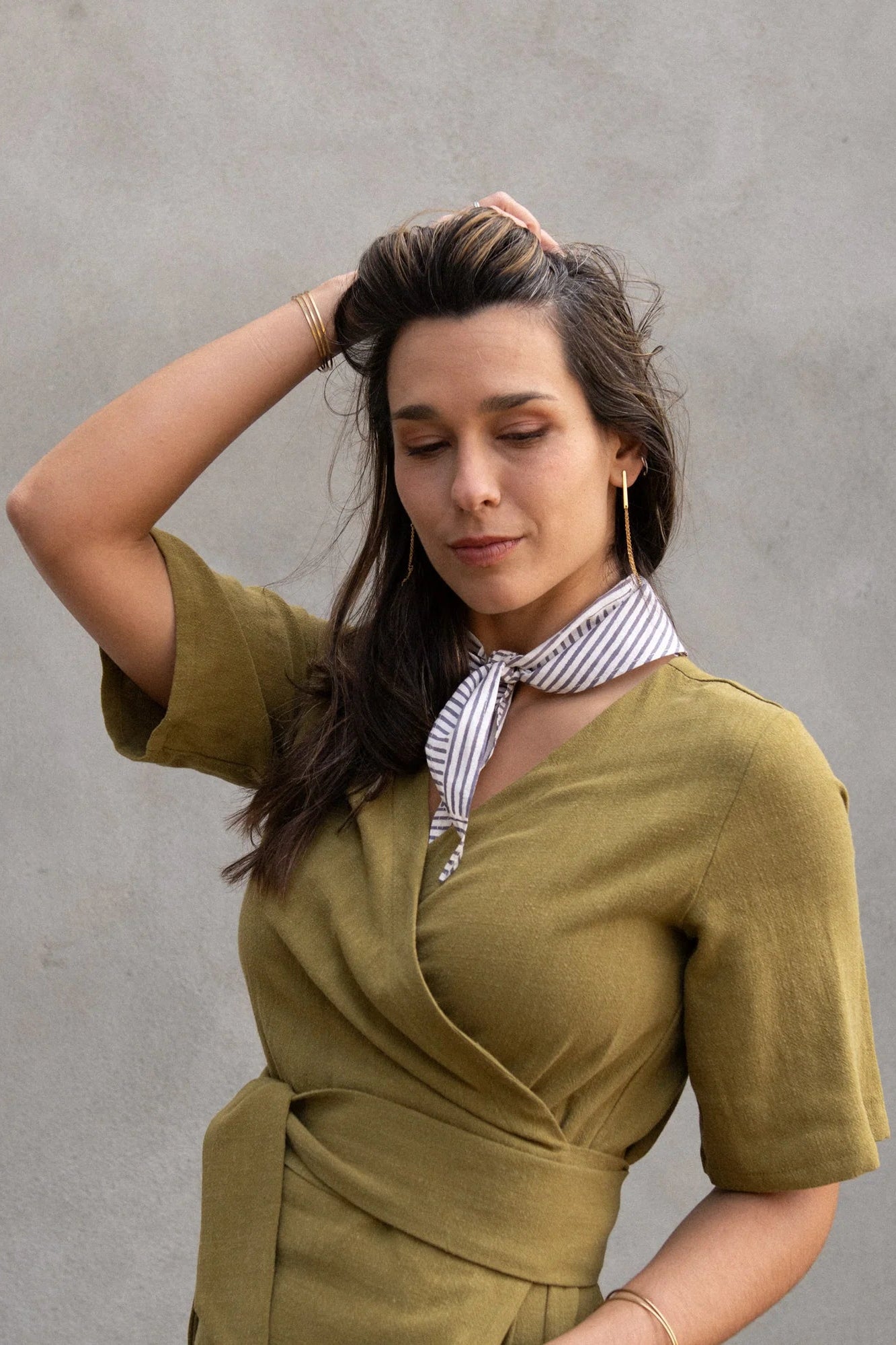 A woman wearing the Helleborus wrap Dress by Kazak in Mousse, standing outside by a white curtain and a cement wall. 