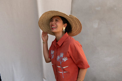 Waist up view of a woman wearing the Hosta Screen-printed Blouse by Kazak in Red with Pysanka, with hand-screen-printed flowers, mid-length raglan sleeves, a classic collar, and a button front. She is wearing it with a matching skirt and straw hat and standing outside by white curtain.