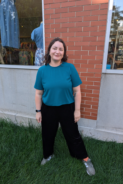 Moira wearing the Flock Exclusive Porto T-shirt by MAS in Teal, standing in front of the brick wall outside of Flock