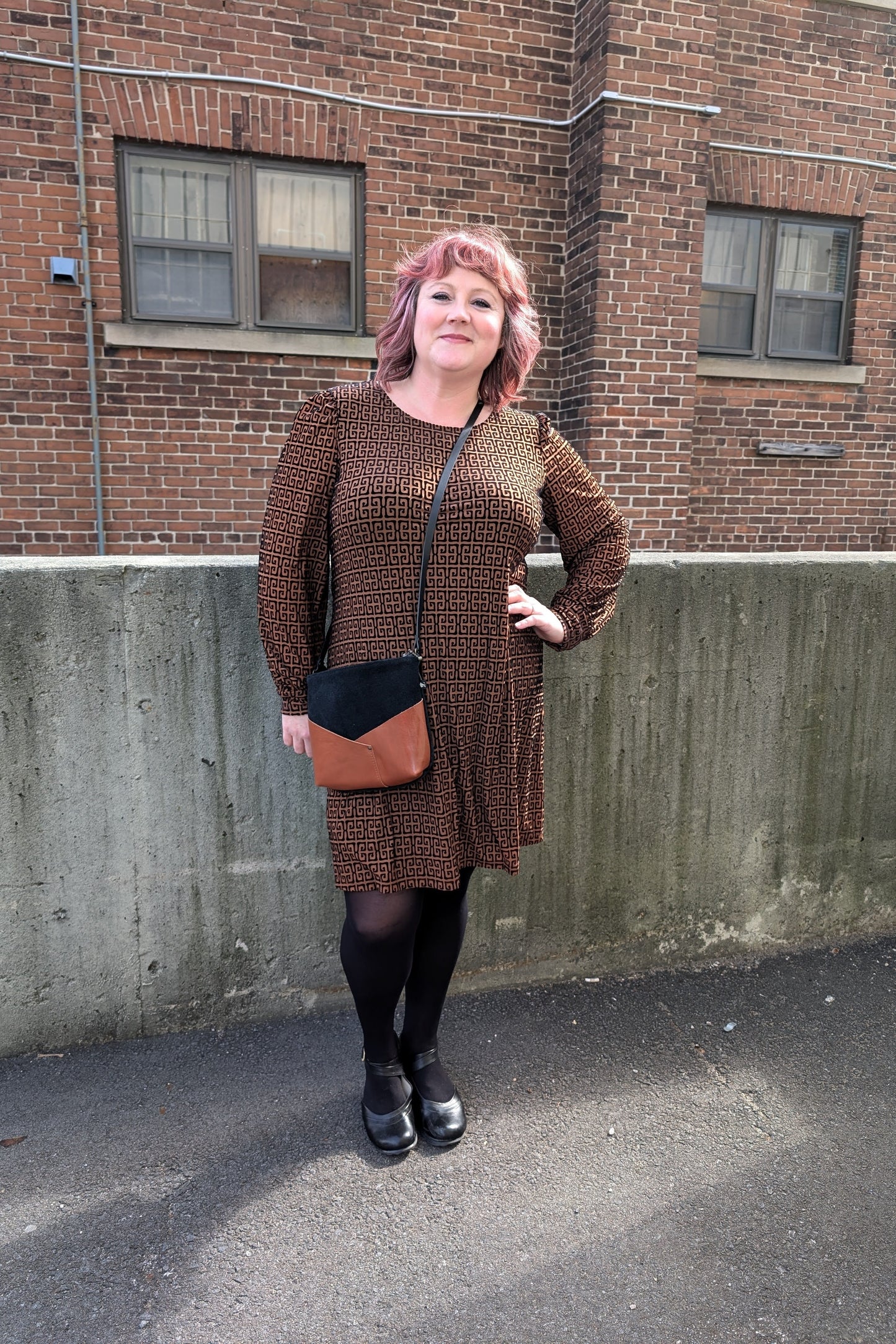 A woman wearing the Gretta Dress by Luc Fontaine in Caramel and Black print, featuring a round neck, slightly puffed sleeves, textured fabric, and above the knee length skirt, standing outside in front of a brick wall. 