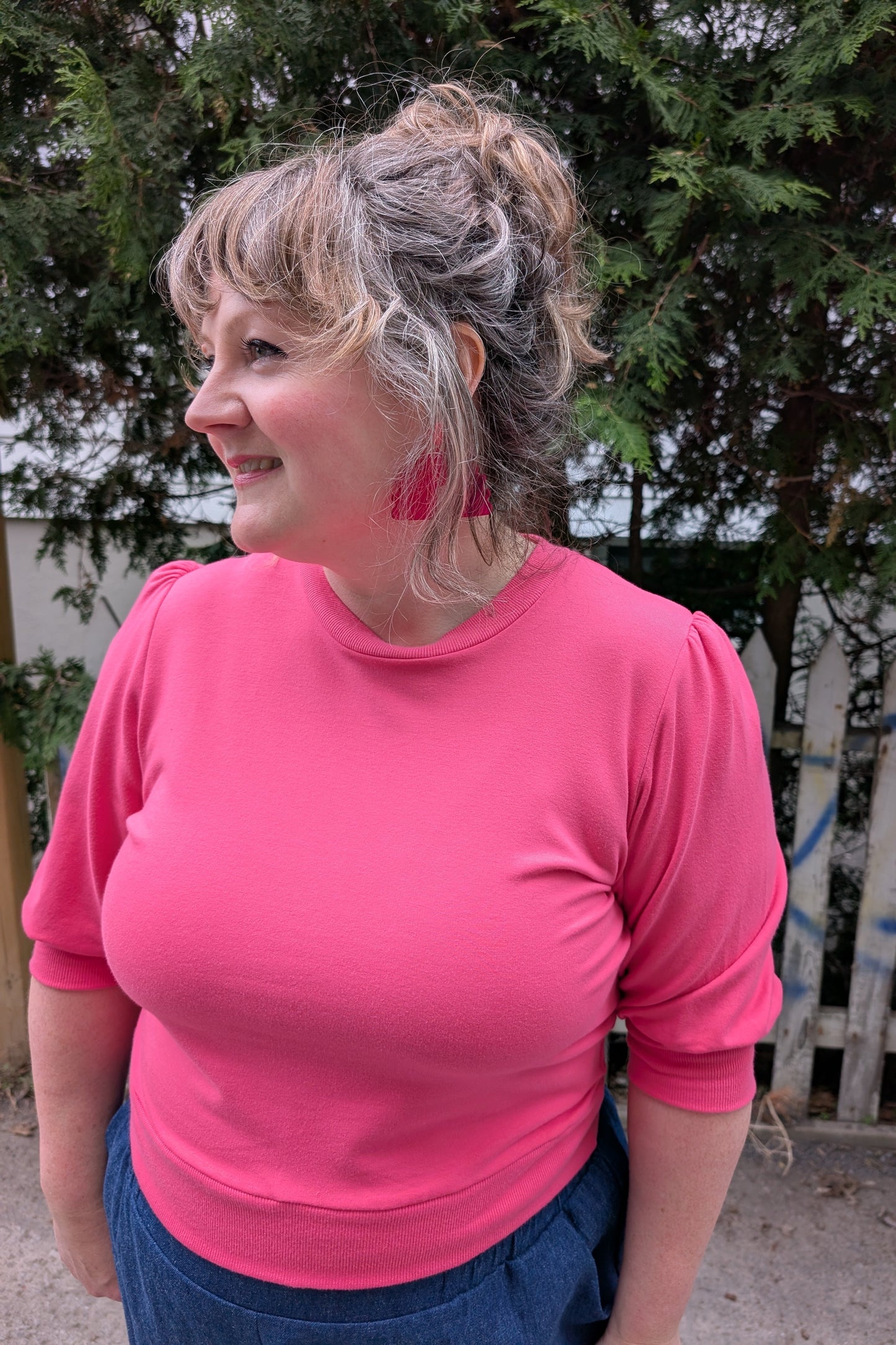 A woman wearing the Finch Sweatshirt by Allison Wonderland in Pink, a cropped sweatshirt with elbow-length puffed sleeves and ribbing at the neck and waistband. She is wearing it with denim pants and standing outside by a graffitied fence and trees.