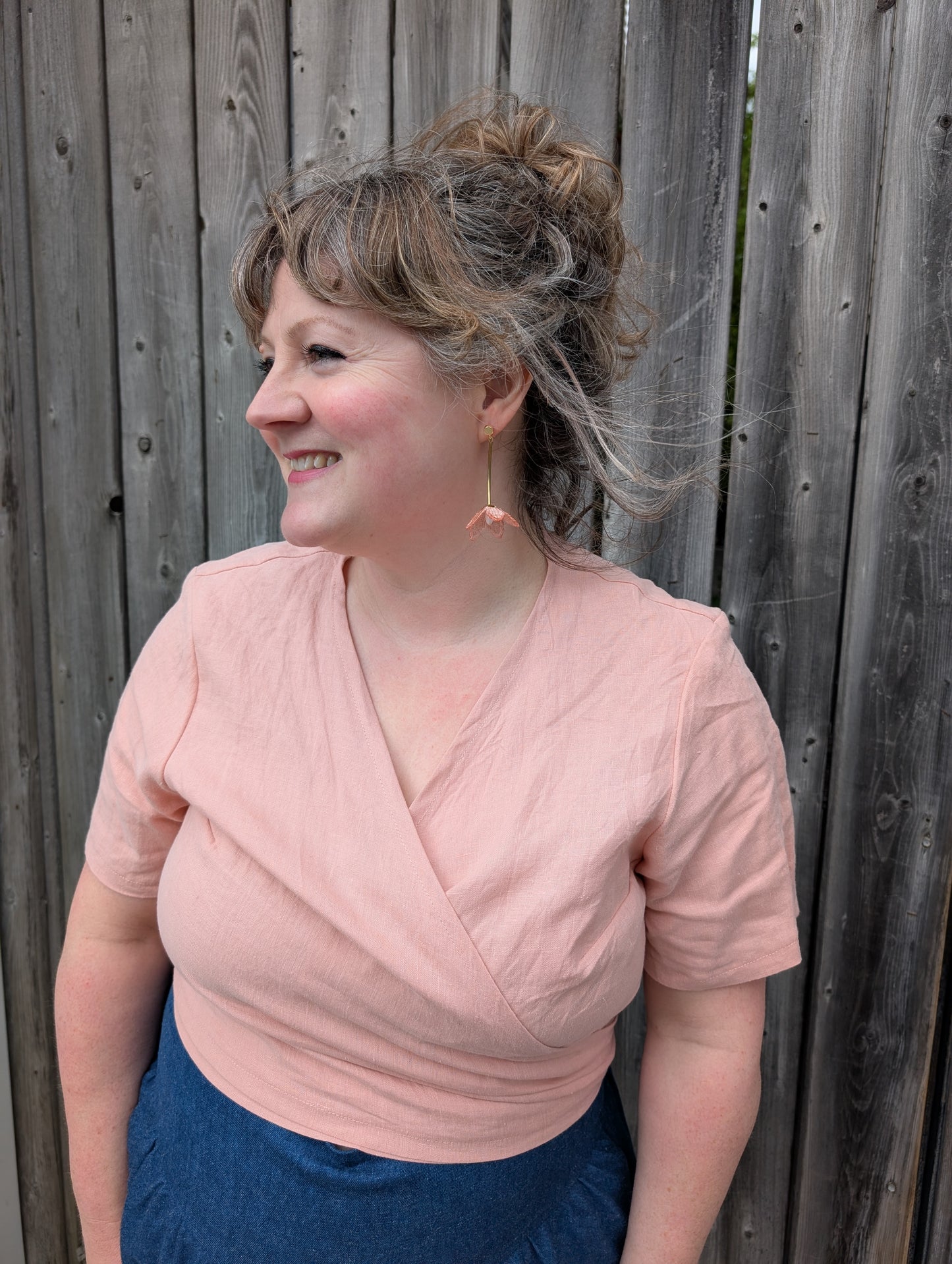 A woman wearing the Stella Top by Lights of All in Ballet Pink, a short sleeved linen wrap top that can be twisted or tied back or front. She is wearing it with the Margot Denim Skirt by Melow and standing by a fence. 