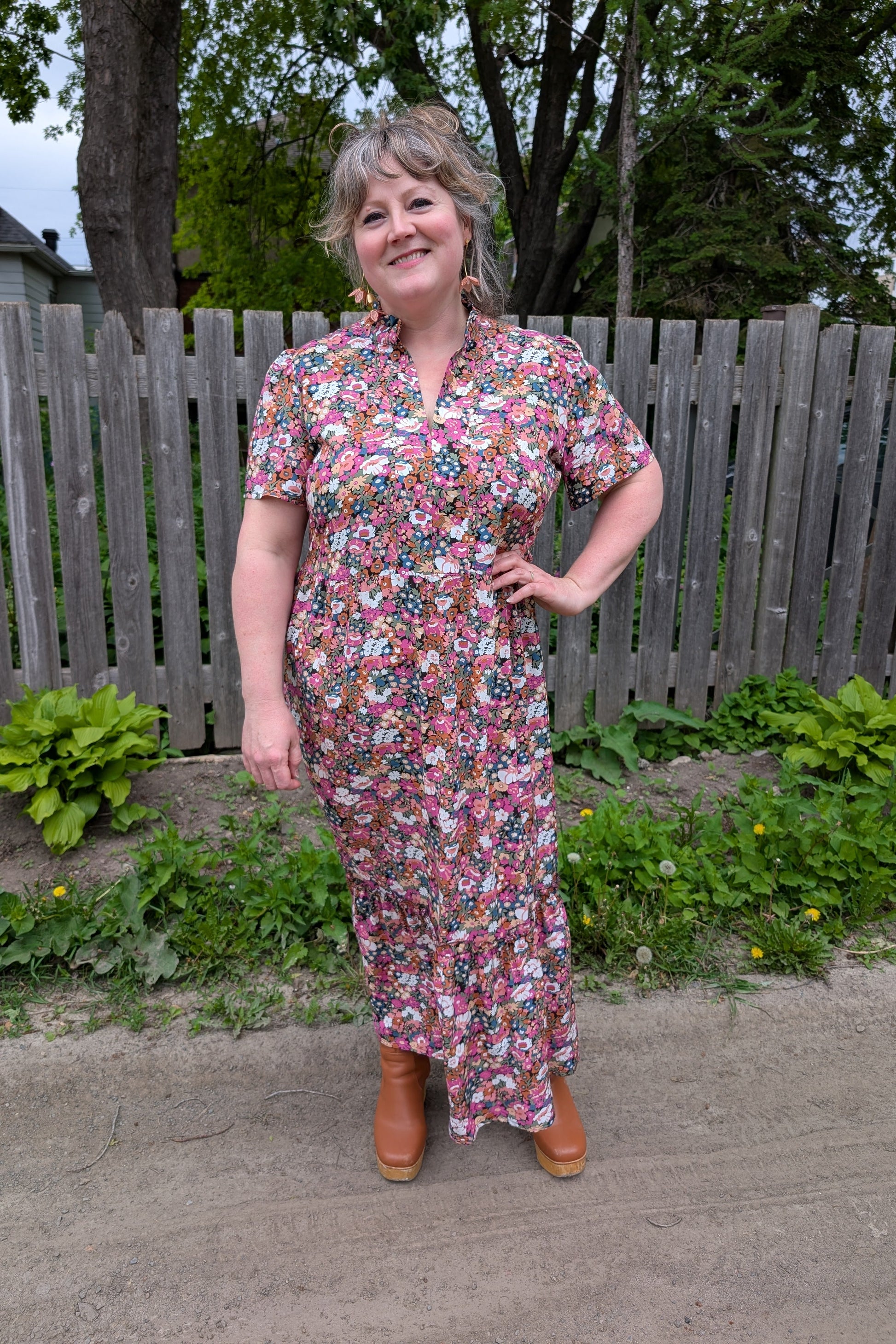 A woman wearing the Petal Print by Dotty in Pink/Blue floral,  a maxi dress with short flutter sleeves, a split V-neck and ruffled collar, with a gathered waist and A-line skirt. She is standing outside by a garden and a wooden fence. 