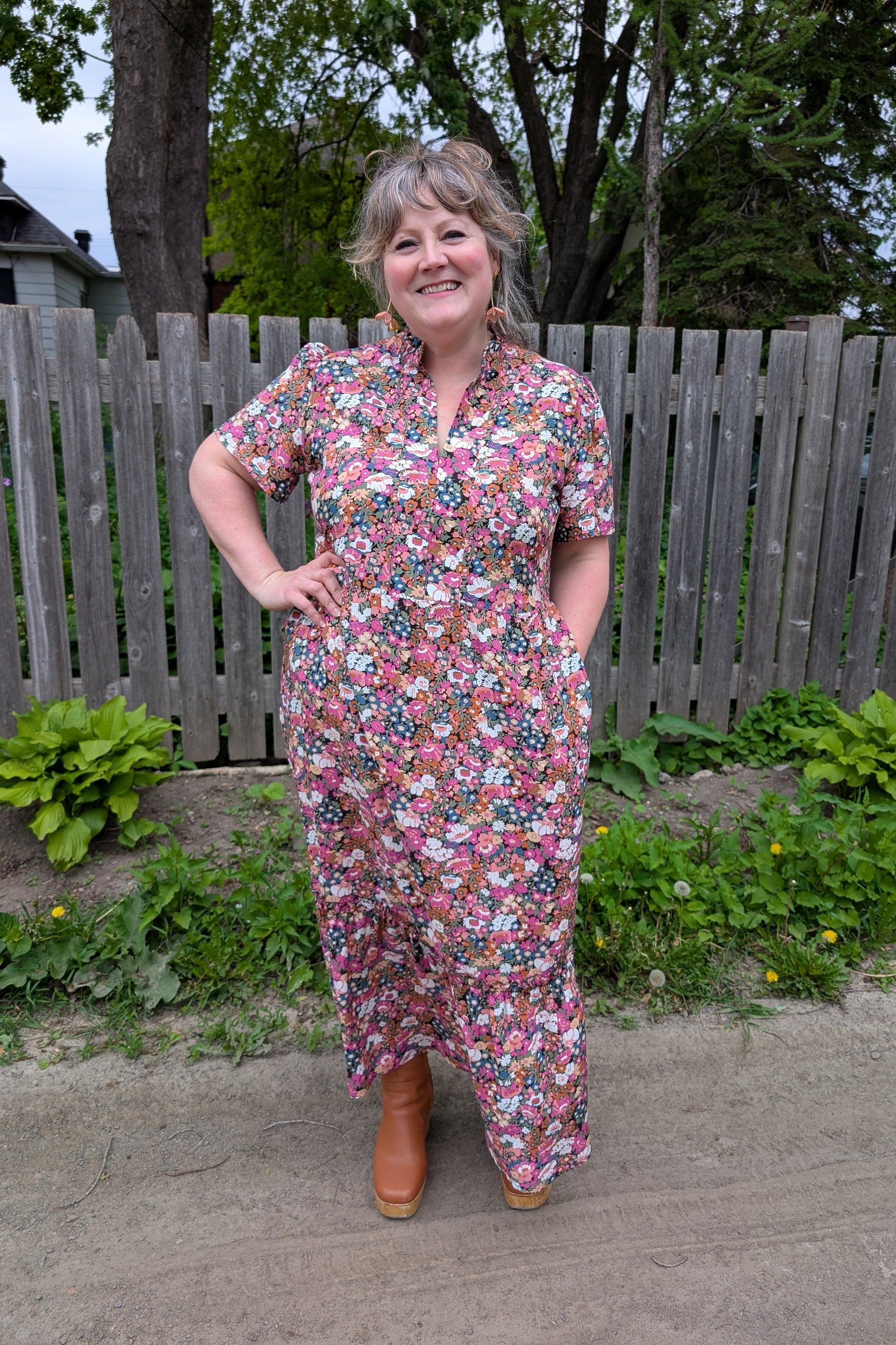 A woman wearing the Petal Print by Dotty in Pink/Blue floral,  a maxi dress with short flutter sleeves, a split V-neck and ruffled collar, with a gathered waist and A-line skirt. She is standing outside by a garden and a wooden fence. 