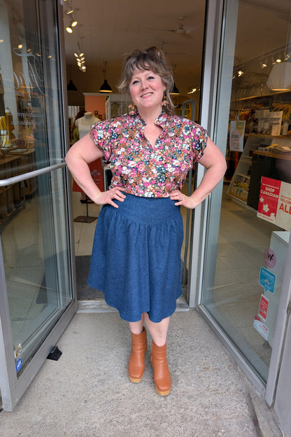 A woman wearing the The Petal Blouse by Dotty in Pink/Blue floral print. It has short floaty sleeves, a split V-neck, a ruffled collar, and a rounded hi-low hemline. She is wearing it with the Margot Skirt by Melow and standing in the doorway at Flock. 