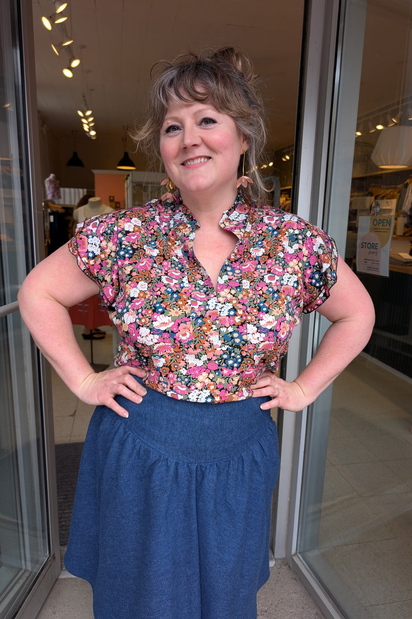 A woman wearing the The Petal Blouse by Dotty in Pink/Blue floral print. It has short floaty sleeves, a split V-neck, a ruffled collar, and a rounded hi-low hemline. She is wearing it with the Margot Skirt by Melow and standing in the doorway at Flock. 