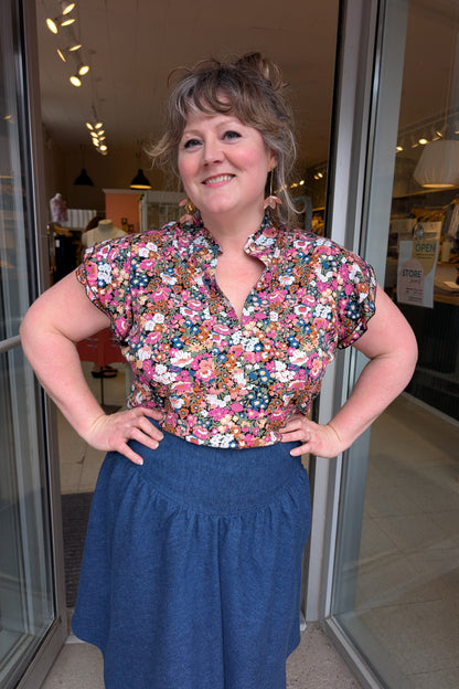 A woman wearing the The Petal Blouse by Dotty in Pink/Blue floral print. It has short floaty sleeves, a split V-neck, a ruffled collar, and a rounded hi-low hemline. She is wearing it with the Margot Skirt by Melow and standing in the doorway at Flock. 