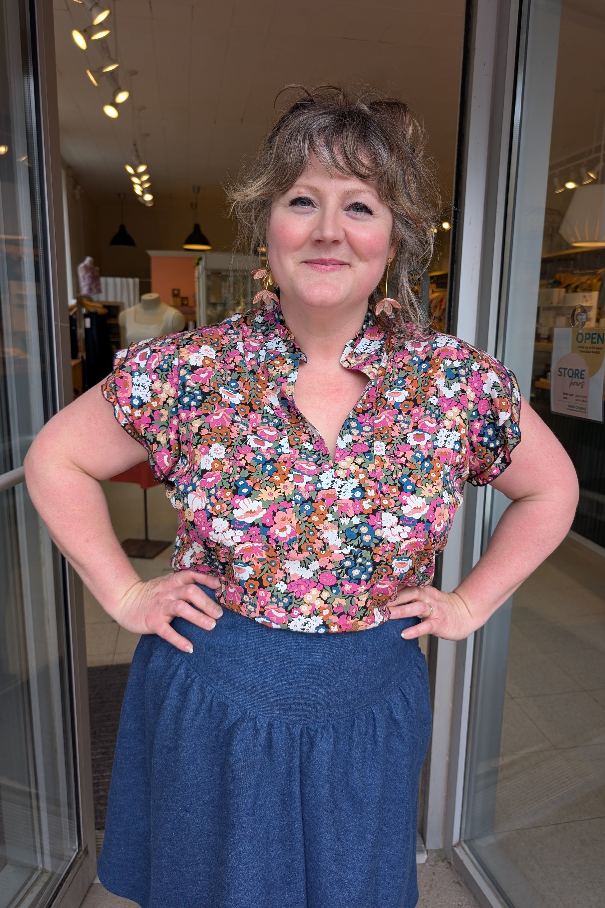 A woman wearing the The Petal Blouse by Dotty in Pink/Blue floral print. It has short floaty sleeves, a split V-neck, a ruffled collar, and a rounded hi-low hemline. She is wearing it with the Margot Skirt by Melow and standing in the doorway at Flock. 
