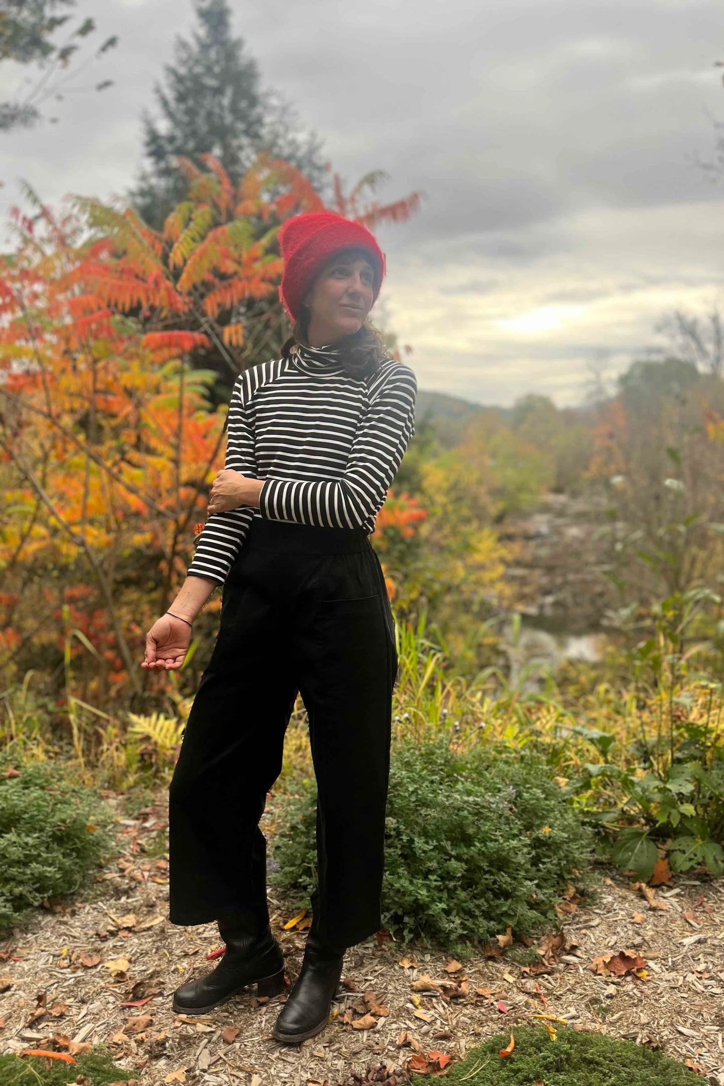 A woman wearing the Olea Pants by 
Kazak in Black, wide-legged ankle-length pants with a flat-front elastic waist and large patch pockets. She is wearing them with the Oris Turtleneck in Black Stripe and standing in a forest.