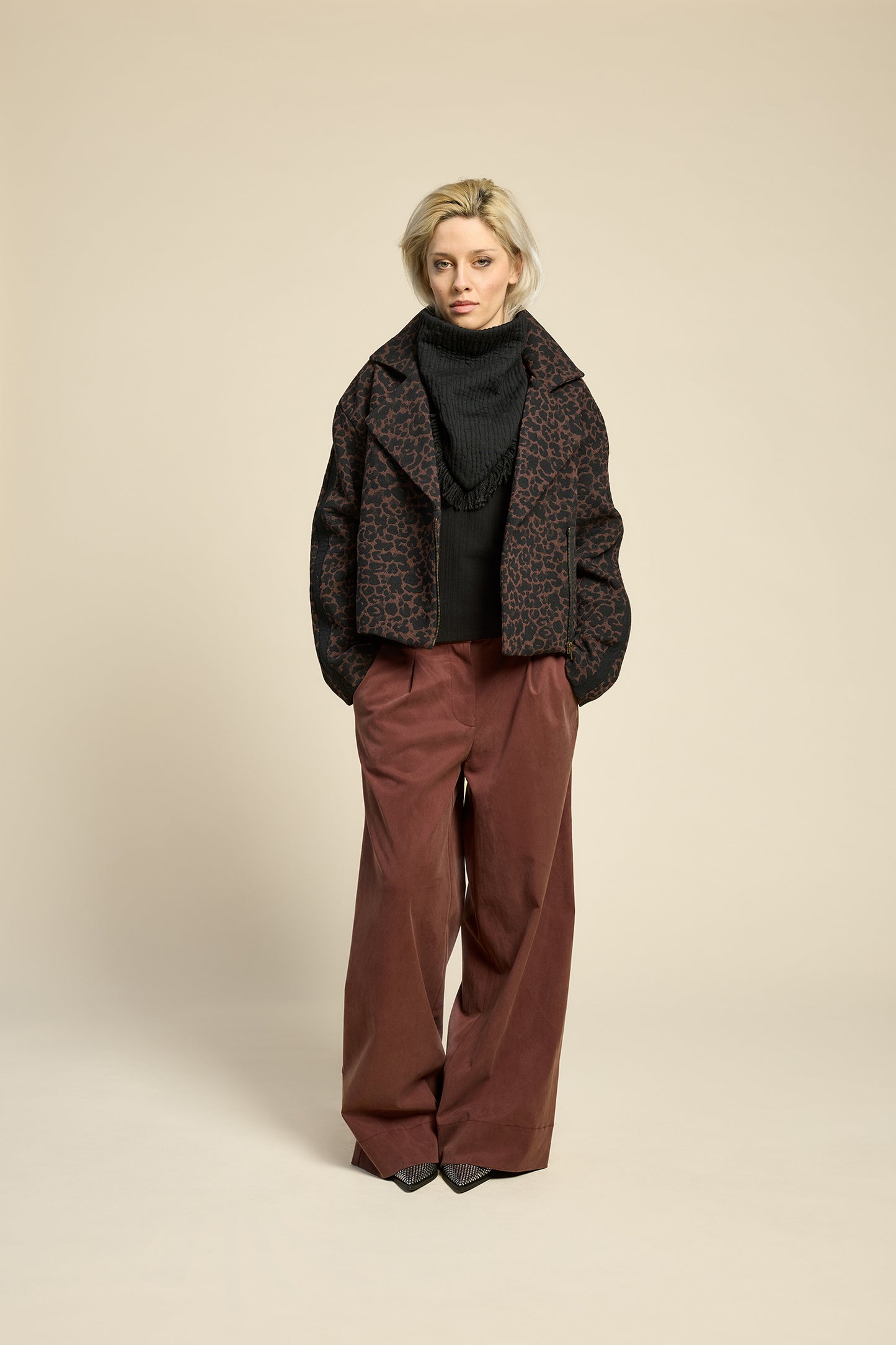 A woman wearing the Perfect Day Jacket by Cokluch in Cappuccino  Leopard, a motorcycle style jacket that's double breasted with a wide collar and zip front. She is wearing it with a black top and brown pants and standing in front of a beige background.