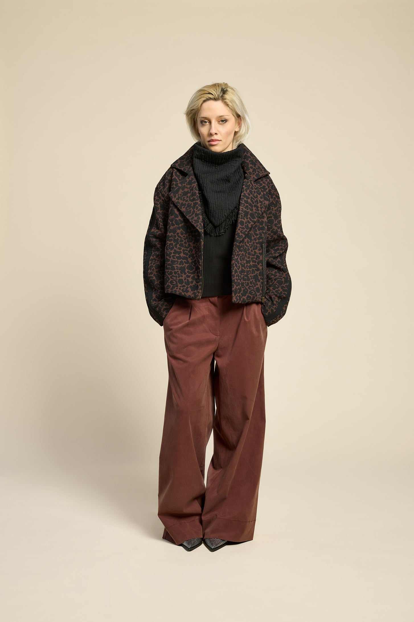 A woman wearing the Perfect Day Jacket by Cokluch in Cappuccino  Leopard, a motorcycle style jacket that's double breasted with a wide collar and zip front. She is wearing it with a black top and brown pants and standing in front of a beige background.