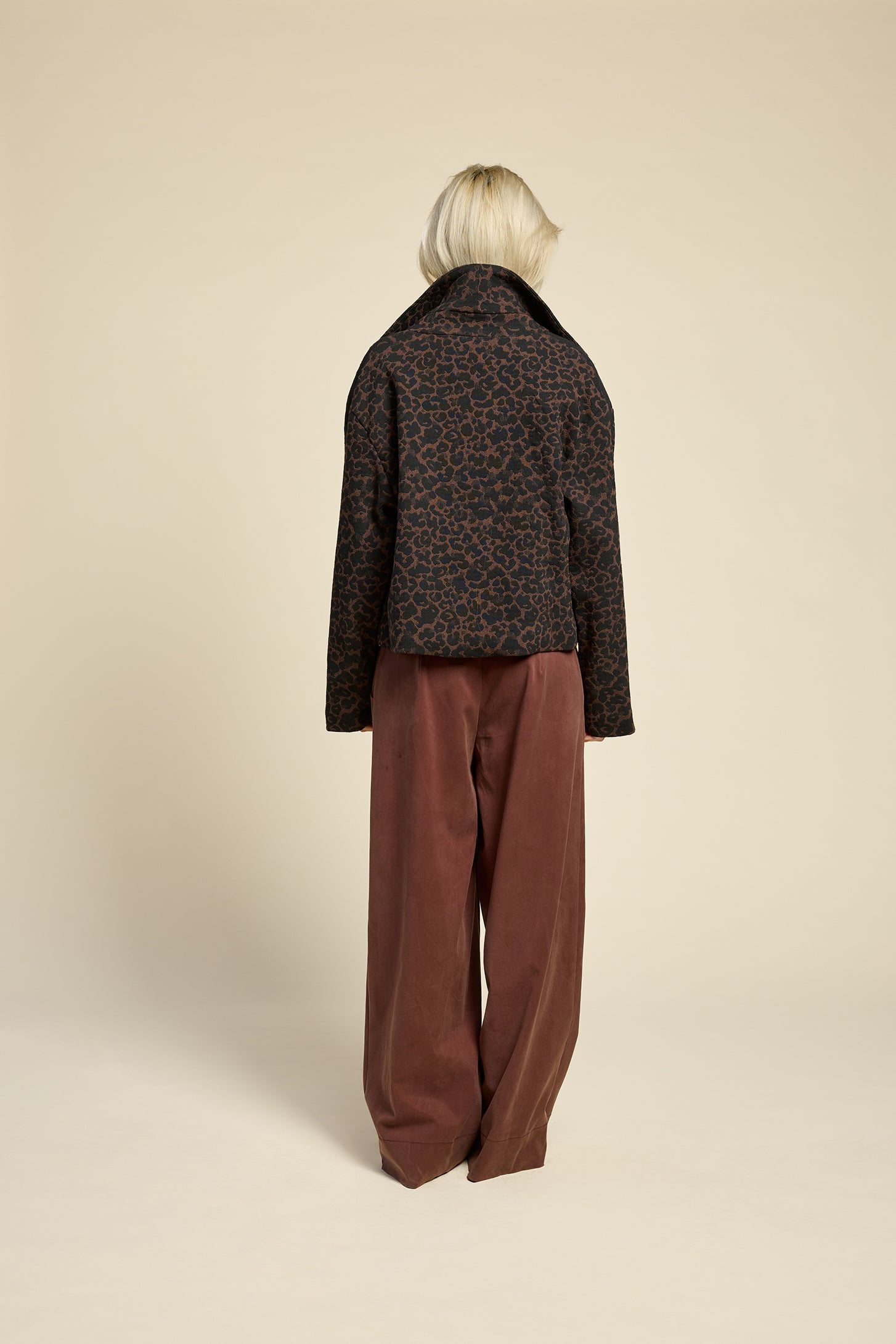 Back view of a woman wearing the Perfect Day Jacket by Cokluch in Cappuccino  Leopard, a motorcycle style jacket that's double breasted with a wide collar and zip front. She is wearing it with a black top and brown pants and standing in front of a beige background.