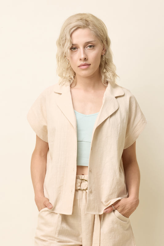 A woman wearing the Phebe Top by Cokluch in Natural, a button front short-sleeved shirt with a rounded blouse collar and a chest pocket. She is wearing it with matching pants and standing in front of a beige background. 