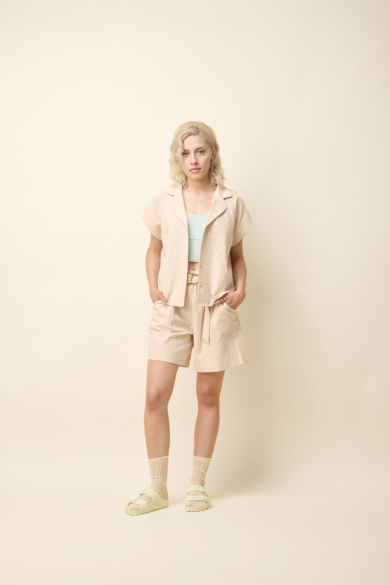 A woman wearing the Phebe Top by Cokluch in Natural, a button front short-sleeved shirt with a rounded blouse collar and a chest pocket. She is wearing it with matching shorts and standing in front of a beige background. 