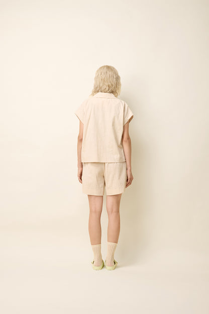 Back view of a woman wearing the Phebe Top by Cokluch in Natural, a button front short-sleeved shirt with a rounded blouse collar and a chest pocket. She is wearing it with matching shorts and standing in front of a beige background. 