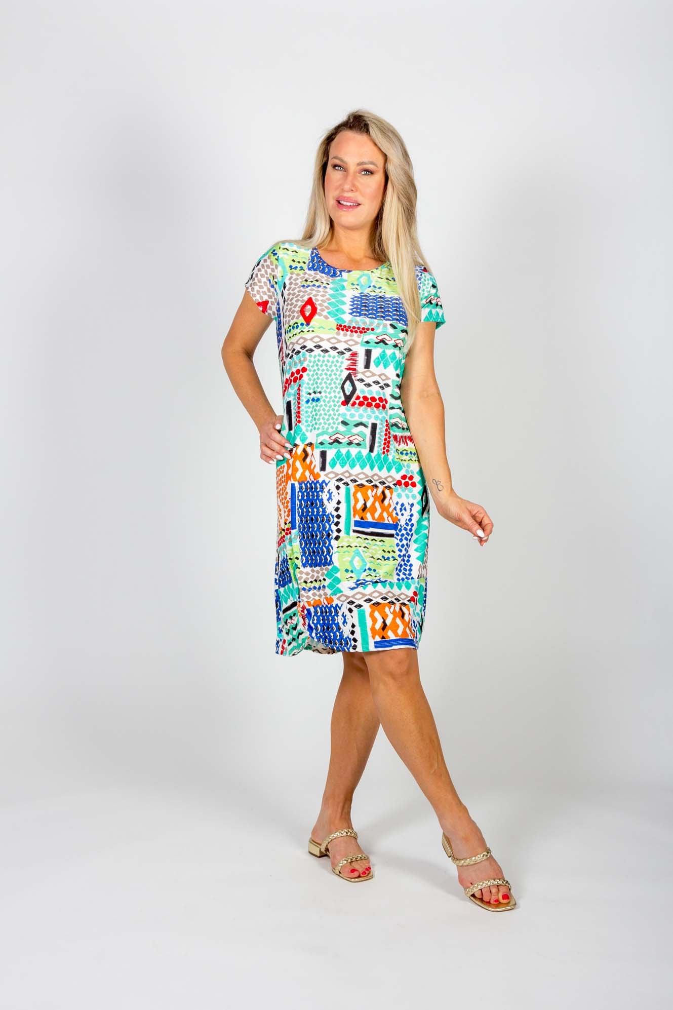 A woman wearing the Chastity Dress by Pure Essence in a geometric Aqua/Blue print stands in front of a white background 