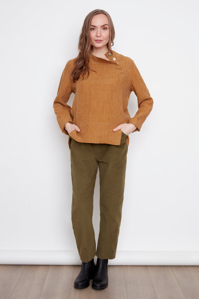A woman wearing the Charlie Top by 
Julie in Ginger Stripes, with an asymmetrical button placket at the neckline and hi-low hemline with side slits. She's wearing them with green pants and standing in front of a white background. 