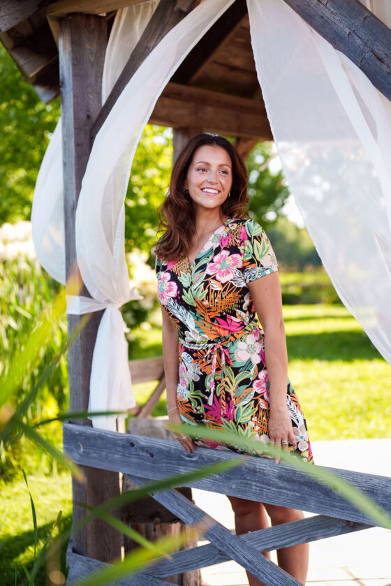 A woman wearing the Macarena Dress by Rien ne se Perd in Tropical print, a short-sleeved faux-wrap dress with a drawstring waist and a flared above-the-knee skirt. She is standing outside in a pagoda.