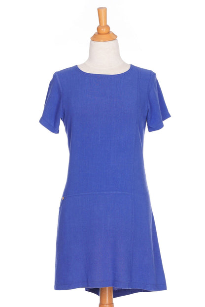 The Merengue Tunic by Rien ne se Perd in Periwinkle, a loose-fitting linen and viscose tunic with a round neck, short sleeves, asymmetrical cut, and patch pocket, is shown on a mannequin in front of a white background. 