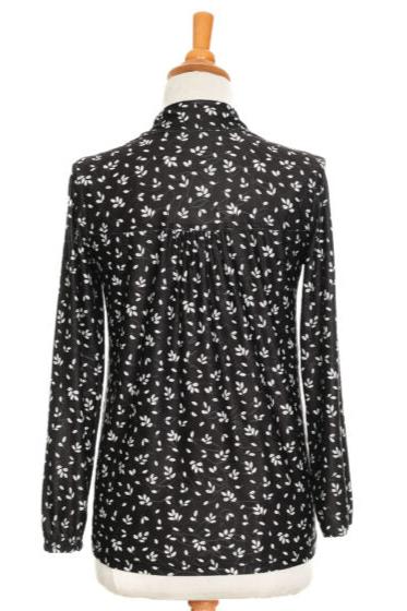 A back view of the Monet Blouse by Rien ne se Perd in Black and White Floral, with a V-neck, faux button placket with coconut buttons, loose fit, and long sleeves with gathered cuffs, is shown on a mannequin in front of a white background 