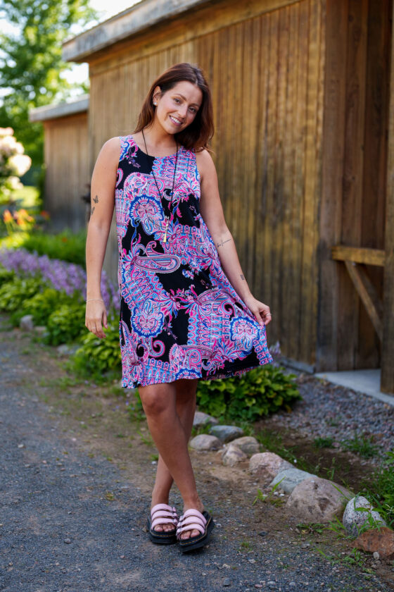 A woman wearing the Musette Dress by Rien ne se Perd, a short sleeveless swing dress with a purple Paisley print. She is standing outside by a wooden building. 