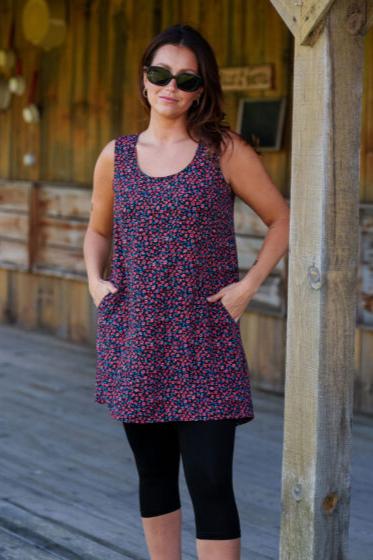 A woman wearing the Oriental Tunic by Rien ne se Perd, featuring a Tiny Flowers print, scoop neck, sleeveless design, trapeze cut, and two pockets. It has a hi-low rounded hemline. She is wearing it with black capri leggings and standing outside in front of a wooden building. 