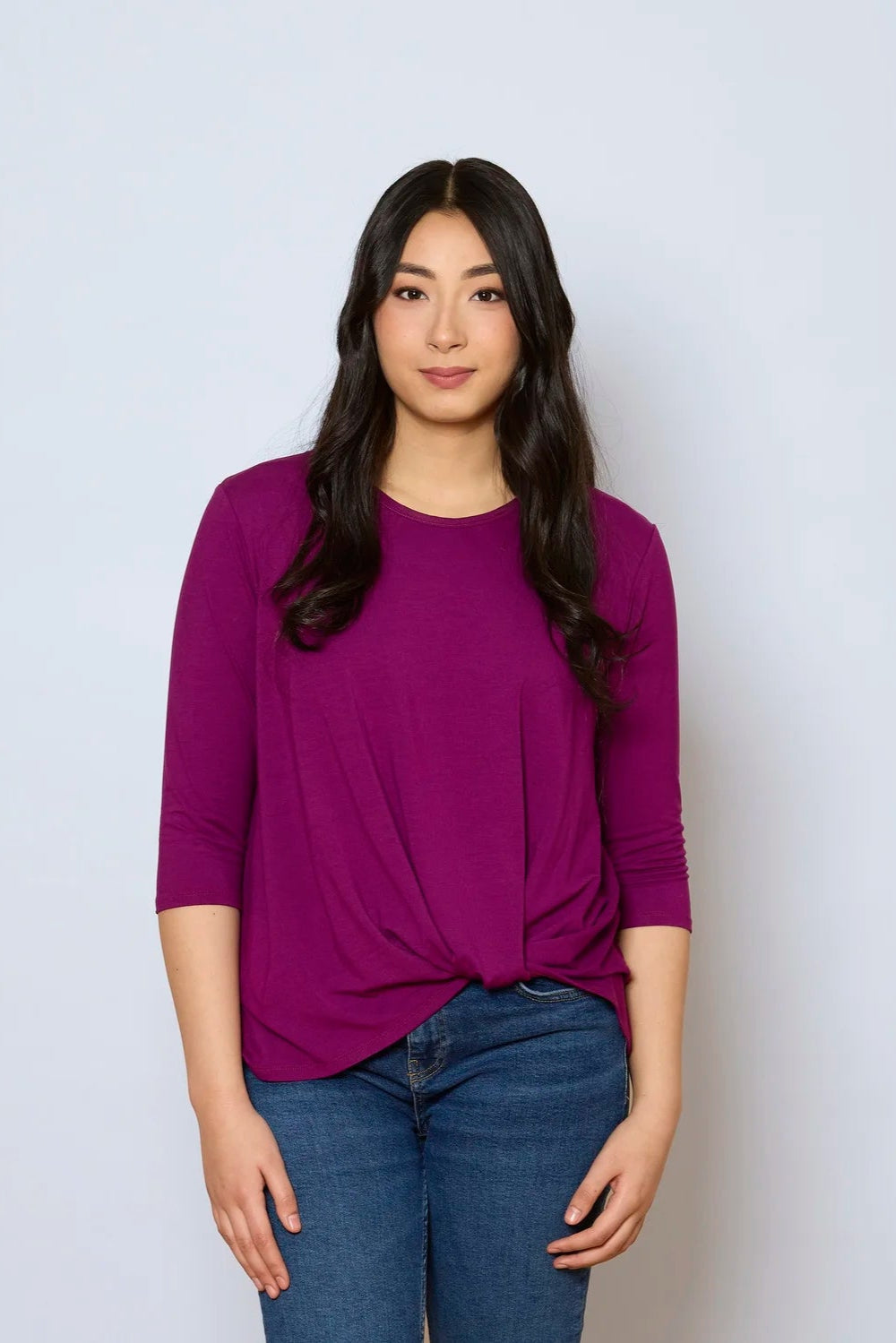 A woman wearing the Reese Top by Tangente in Fuchsia, with a round neck, 3/4 drop sleeves, and a twisted hem in front. She is wearing it with jeans and standing in front of a white background. 