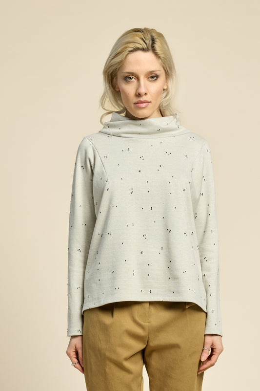 Waist up view of a woman wearing the Reminder Sweater by Cokluch in Natural, with a high twisted neck, darts and the shoulders, long sleeves, and a loose fit. She is wearing it with yellow pants and standing in front of a beige background. 